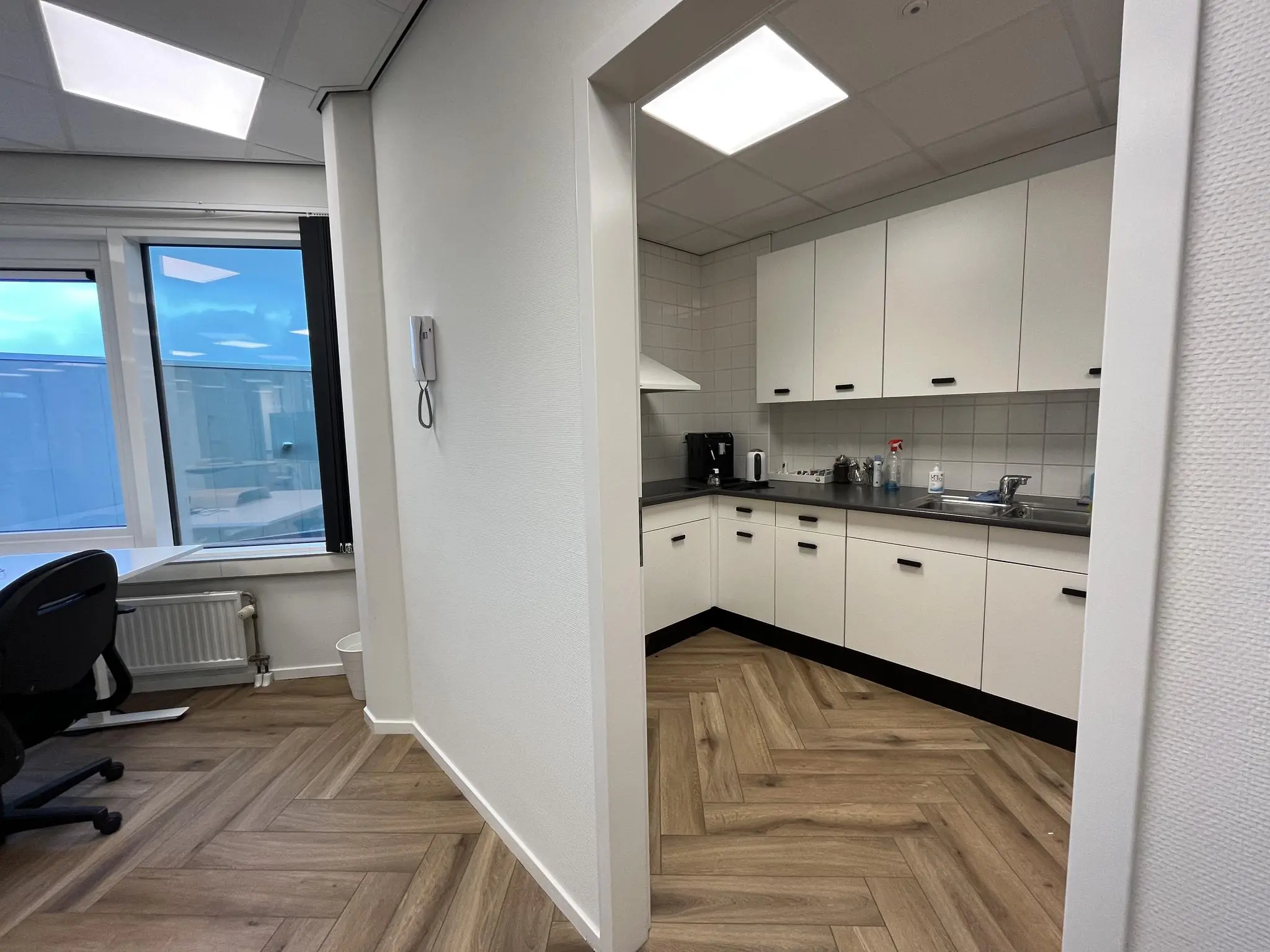 Kleine kantoorkeuken met witte kasten en visgraat pvc-vloer aan de Lange Dreef.